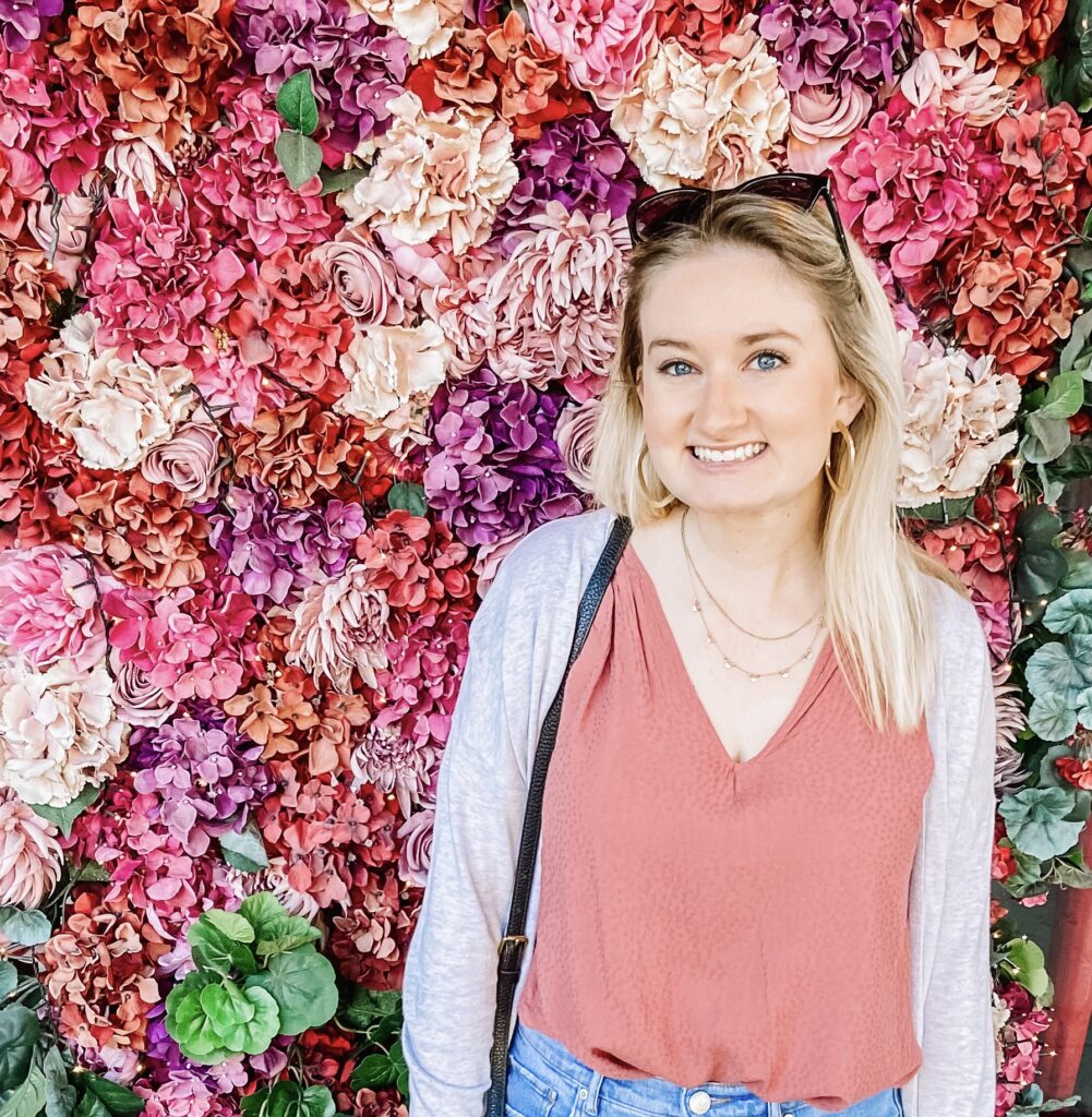 profesora rubia parada frente a la pared de flores sonriendo