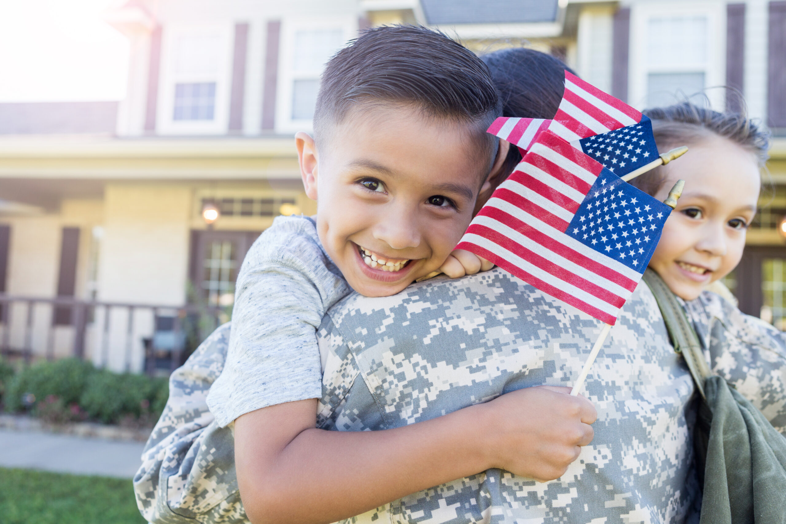 young boy smiles as he holds a flag and hugs his mother in uniform